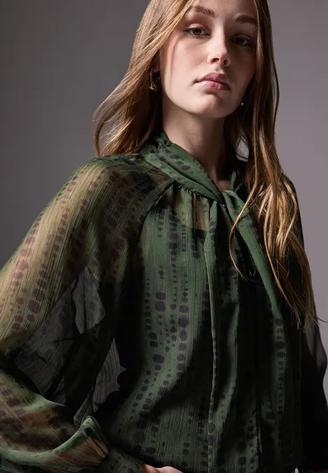 Frau mit langen Haaren trägt eine transparente, grüne Bluse mit Muster.